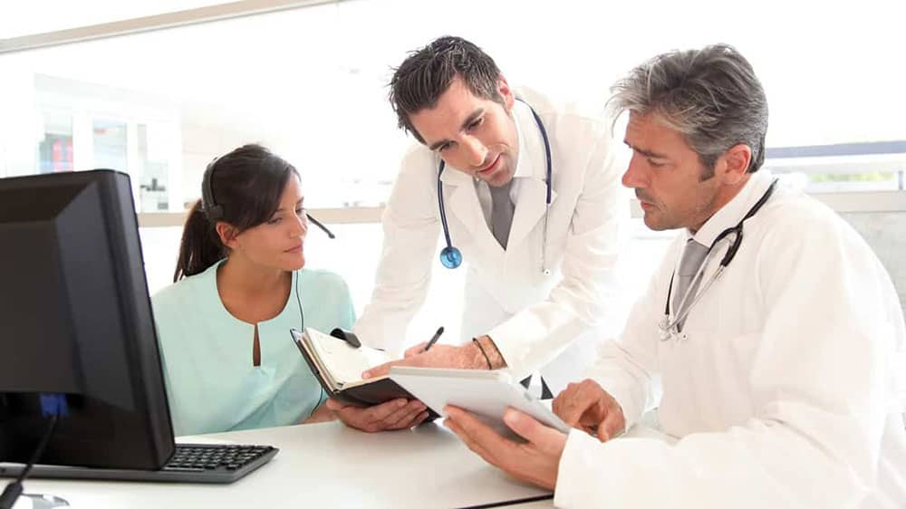 Female in a green top, with a man in a white coat, and another man standing between them looking at patient files.