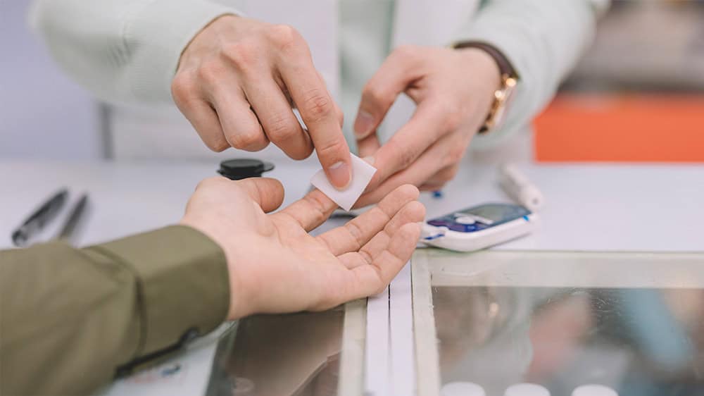 A health professional wipes the inside of a finger of a man with a bgl meter and lancet device on the table in the background.