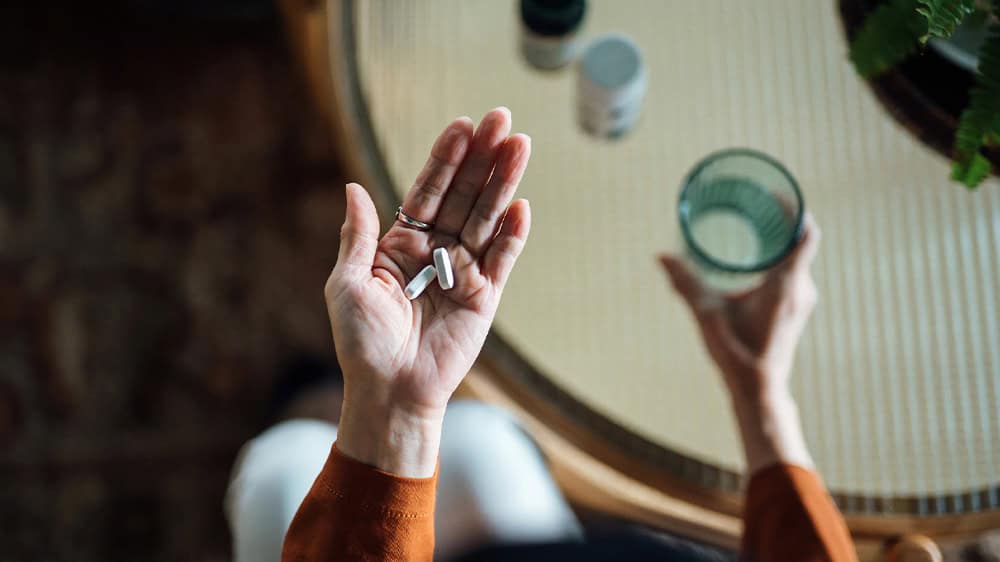 Overhead picture of a woman holding pills in one hand and a glass of water in the other with other medications on a table under her hand.